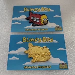 Puede incluir: Dos tarjetas con un fondo de cielo azul y nubes blancas. Cada tarjeta presenta un pin de esmalte dorado y rojo de Homer Simpson en un dirigible. El dirigible está etiquetado como "Blimpy Boy" y el texto "@bakterstockpins" está en cada tarjeta.