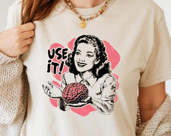 May include: Beige t-shirt with a retro graphic of a woman holding a brain, with the text "USE IT!" in black and pink. The design is set against a pink heart-shaped background. The shirt is a casual, everyday wear item.