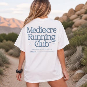 May include: White t-shirt featuring the text "Mediocre Running Club" in blue. The shirt also includes the text "ESTD. 1891" and "Enjoying every step at our own pace". The t-shirt has a relaxed fit and short sleeves.
