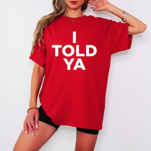 May include: A red t-shirt with the text "I TOLD YA" in large, white, block letters. The shirt is a solid colour and appears to be made of a soft material. The shirt is being worn.