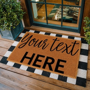 May include: A rectangular natural coir doormat with black text reading Your text HERE, layered over a larger black and white buffalo check rug. The doormat features a coarse, fibrous texture and sits on a wooden porch floor in front of a door.
