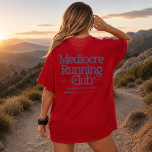 A woman wearing a red t-shirt with the text "Mediore Running Club" standing on a dirt path in a mountainous landscape at sunset.