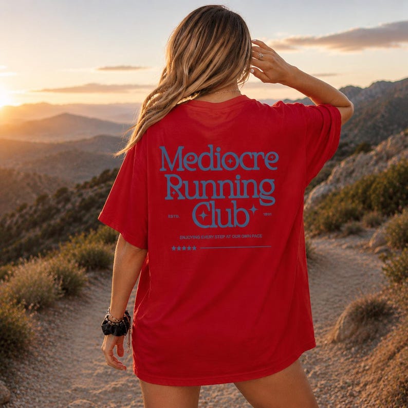A woman wearing a red t-shirt with the text "Mediore Running Club" standing on a dirt path in a mountainous landscape at sunset.