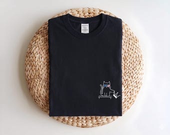 May include: A black crew neck t-shirt featuring a white line art graphic of a cat wearing red and blue 3D glasses, sitting on a bench and holding a crown. The shirt has a white neck label with black text and is folded neatly.