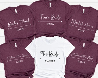 May include: Five maroon and white t-shirts with text that reads "Brides Maid", "Team Bride", "Maid of Honor", "Mother of the Groom", and "Mother of the Bride". Each shirt has a name below the text. The names are Daisy, Daisy, Kate, Asley, and Sally.