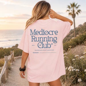 A woman wearing a pink t-shirt with the text "Mediocre Running Club" standing on a beach with palm trees and a sunset in the background.