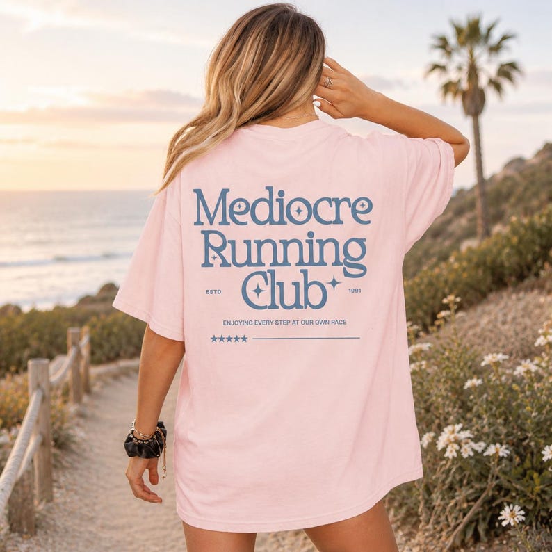 A woman wearing a pink t-shirt with the text "Mediocre Running Club" standing on a beach with palm trees and a sunset in the background.
