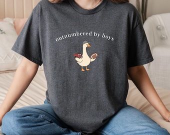 May include: Dark gray t-shirt with the text "outnumbered by boys." The shirt features a cartoon goose wearing a hat and holding a football and a toy car. The shirt is made of a soft material.