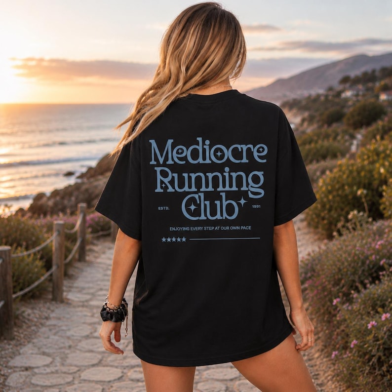A woman wearing a black t-shirt with text on the back is standing on a path near the ocean, with a sunset in the background.