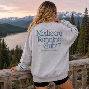 A woman wearing a gray sweatshirt with the text "Mediocre Running Club" on the back, standing on a wooden deck overlooking a scenic mountain landscape.