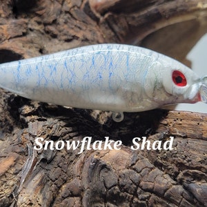 May include: A white fishing lure with blue and silver accents. The lure has a red eye and a clear plastic bill. The lure is on a piece of wood with the text "Snowflake Shad" written below it.