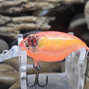 May include: A bright orange fishing lure with a yellow crackle pattern, a clear plastic diving lip, and a prominent red eye. The lure features two metal treble hooks attached to the underside and rear, designed for catching fish in water.