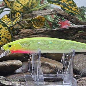 May include: A neon yellow and white fishing lure with a glitter finish, a pink accent near the head, a clear plastic diving lip, two silver treble hooks, and a white feathered tail hook, mounted on a clear plastic display stand.