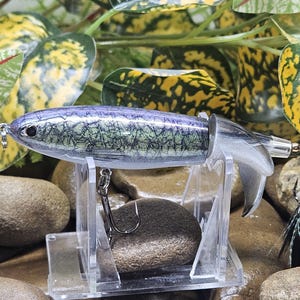 May include: A fishing lure with a metallic purple and silver body featuring a textured scale pattern. The lure has a silver tail section, a metal hook attached to the underside, and a rear hook adorned with black and white feathers and a red band.