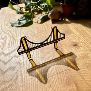 Puede incluir: Un adorno de vidrio tintado que representa el Puente Golden Gate en negro y dorado. El adorno est&aacute; colgado de una cuerda y proyecta una sombra sobre una superficie de madera.