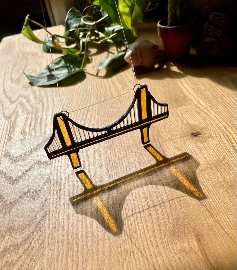 Puede incluir: Un adorno de vidrio tintado que representa el Puente Golden Gate en negro y dorado. El adorno est&aacute; colgado de una cuerda y proyecta una sombra sobre una superficie de madera.
