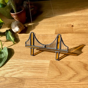 Puede incluir: Una representaci&oacute;n en vitral del Puente Golden Gate, con un esquema de colores amarillo y negro. El puente est&aacute; suspendido de una cuerda y cuelga sobre un fondo de madera.