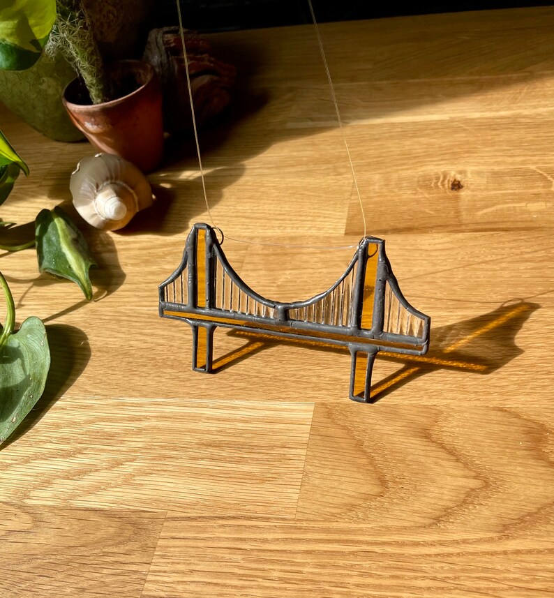Puede incluir: Una representaci&oacute;n en vitral del Puente Golden Gate, con un esquema de colores amarillo y negro. El puente est&aacute; suspendido de una cuerda y cuelga sobre un fondo de madera.
