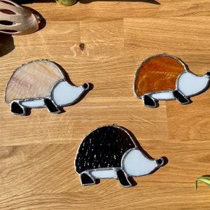 May include: Three stained glass hedgehogs, one white, one brown, and one black, with white faces and black eyes. The hedgehogs are on a wooden surface.