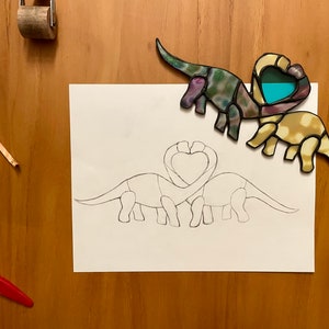 May include: Two stained glass dinosaurs in the shape of a heart, with one dinosaur in yellow and the other in purple and green, are placed on a white sheet of paper with a pencil drawing of two dinosaurs in the shape of a heart.