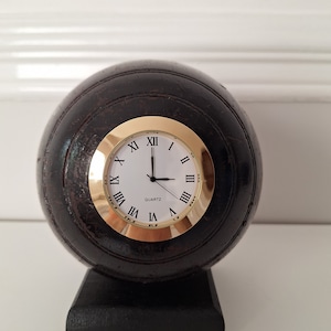 May include: A brown wooden clock shaped like a bowling ball with a gold rim around the white face. The clock face has Roman numerals and the word "QUARTZ" printed below the 12.