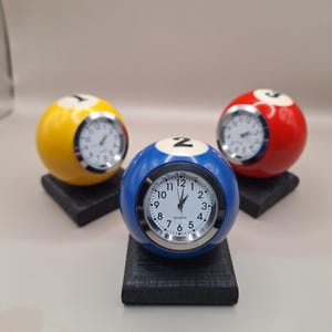 May include: Three spherical quartz clocks designed as billiard balls in yellow, blue, and red, each featuring a white face with black numerals and hands. Each clock is mounted on a small, square, black textured base for stability.