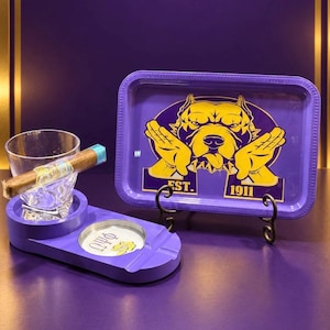 May include: A purple rectangular rolling tray featuring a yellow pit bull graphic with hands and the text EST. 1911, paired with a purple cigar ashtray stand holding a clear glass and a cigar, with the Greek letters Omega Psi Phi printed on the base.