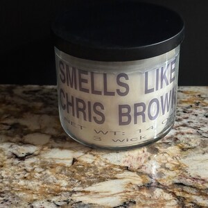 Puede incluir: Un tarro de vela blanco con tapa negra. El tarro tiene una etiqueta que dice "Smells Like Chris Brown" y "Net Wt: 14 oz 3 Wick".