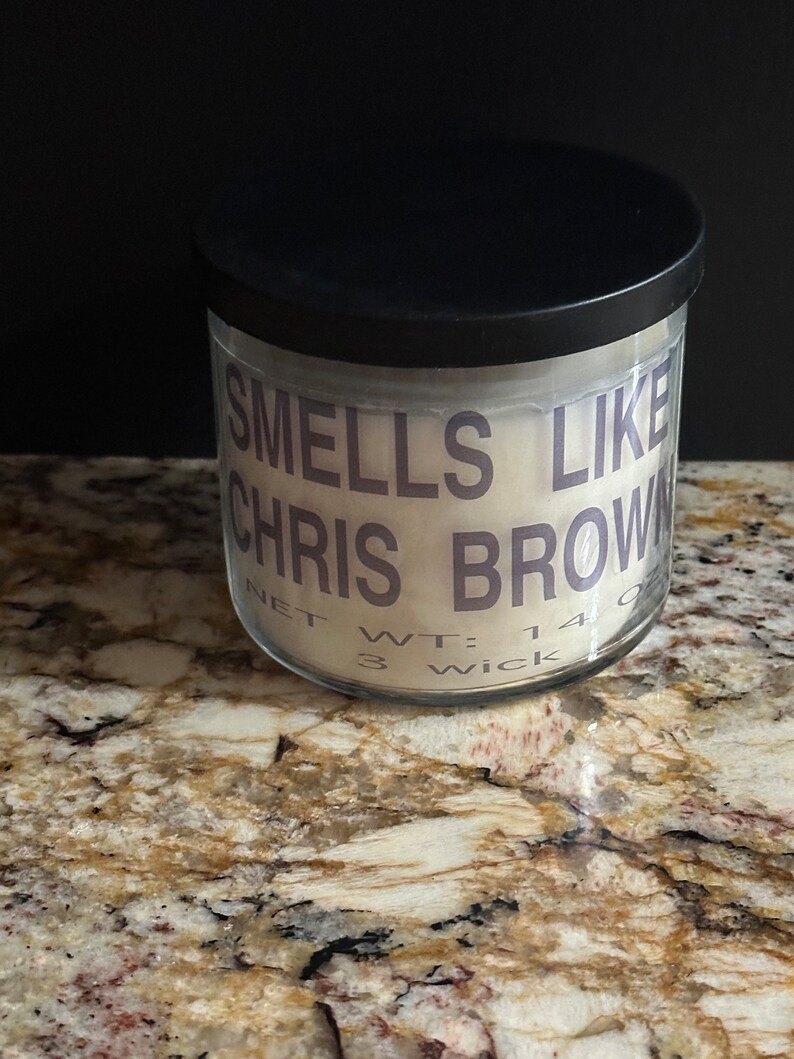 Puede incluir: Un tarro de vela blanco con tapa negra. El tarro tiene una etiqueta que dice "Smells Like Chris Brown" y "Net Wt: 14 oz 3 Wick".