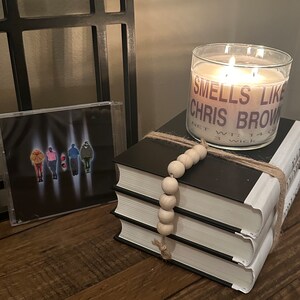 Puede incluir: Una vela blanca con una etiqueta negra que dice "Smells Like Chris Brown" y "Net Wt: 14 oz 3 Wick" est&aacute; encendida y colocada sobre una pila de tres libros. Los libros est&aacute;n envueltos con cuerda y una cadena de cuentas de madera.