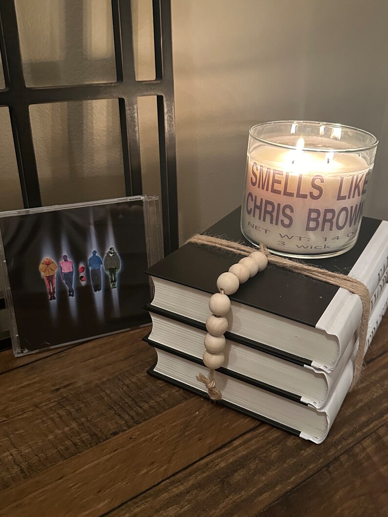 Puede incluir: Una vela blanca con una etiqueta negra que dice "Smells Like Chris Brown" y "Net Wt: 14 oz 3 Wick" est&aacute; encendida y colocada sobre una pila de tres libros. Los libros est&aacute;n envueltos con cuerda y una cadena de cuentas de madera.