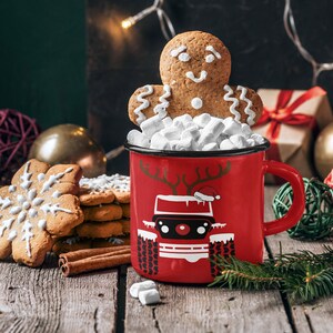 Puede incluir: Una taza roja con un hombre de jengibre y malvaviscos en la parte superior. La taza tiene un dise&ntilde;o blanco de un coche con cuernos de reno y un gorro de Pap&aacute; Noel. La taza est&aacute; sobre una mesa de madera con galletas de jengibre y palitos de canela.