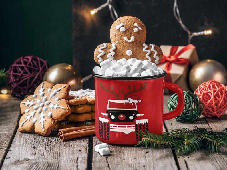 Puede incluir: Una taza roja con un hombre de jengibre y malvaviscos en la parte superior. La taza tiene un dise&ntilde;o blanco de un coche con cuernos de reno y un gorro de Pap&aacute; Noel. La taza est&aacute; sobre una mesa de madera con galletas de jengibre y palitos de canela.