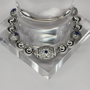 May include: A silver bracelet with a football charm. The football charm is decorated with white rhinestones and a blue star. The bracelet is made of silver beads with rhinestones.