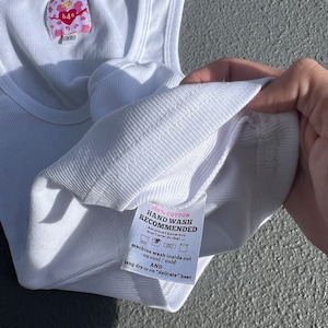 May include: White ribbed tank top with a pink and white label that says "h40" and "XS". The label also includes instructions for washing the tank top: "100% Cotton, Hand Wash Recommended, but since I know you won't ever do that... machine wash inside out on cool/cold AND hang dry or on "delicate" heat"