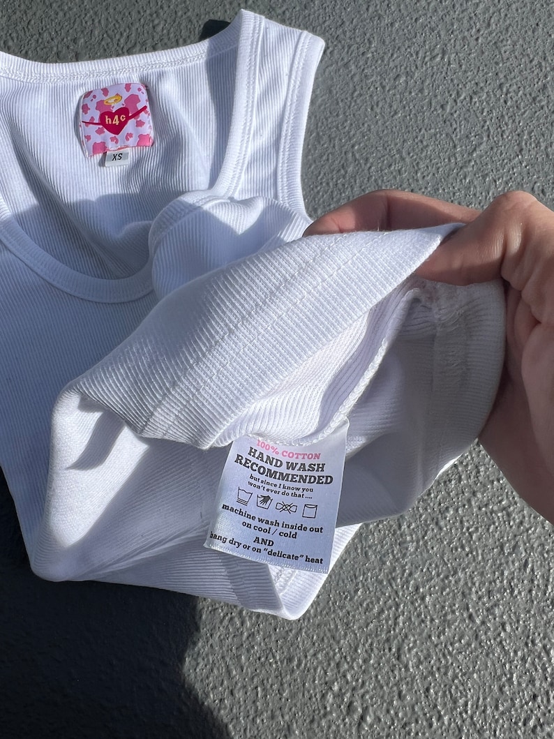 May include: White ribbed tank top with a pink and white label that says "h40" and "XS". The label also includes instructions for washing the tank top: "100% Cotton, Hand Wash Recommended, but since I know you won't ever do that... machine wash inside out on cool/cold AND hang dry or on "delicate" heat"