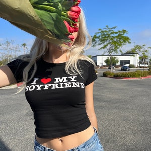 May include: A woman wearing a black crop top with the text "I love my boyfriend" in white letters and a red heart. She is wearing blue jeans and has a belly button ring. She is holding a bouquet of red tulips in front of her face.
