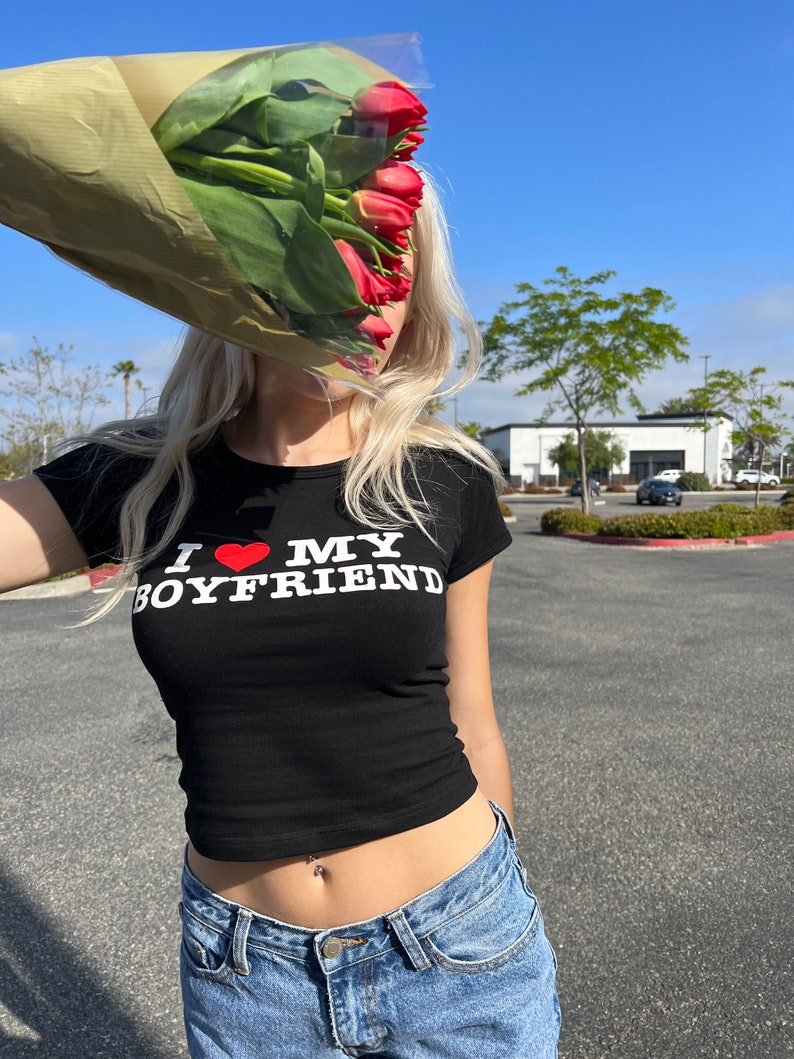 May include: A woman wearing a black crop top with the text "I love my boyfriend" in white letters and a red heart. She is wearing blue jeans and has a belly button ring. She is holding a bouquet of red tulips in front of her face.