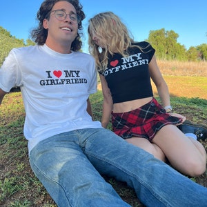 May include: A couple wearing matching shirts with the text "I love my girlfriend" and "I love my boyfriend" on them. The woman is wearing a black crop top with a red heart and a red and black plaid skirt. The man is wearing a white t-shirt with a red heart and blue jeans.