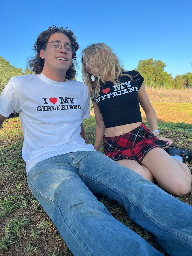 May include: A couple wearing matching shirts with the text "I love my girlfriend" and "I love my boyfriend" on them. The woman is wearing a black crop top with a red heart and a red and black plaid skirt. The man is wearing a white t-shirt with a red heart and blue jeans.