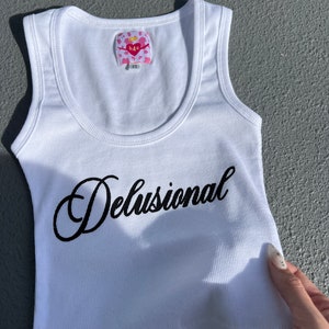 May include: White ribbed tank top with a black cursive inscription that reads "Delusional". The tank top has a pink and white label that reads "h4o" with a heart symbol.