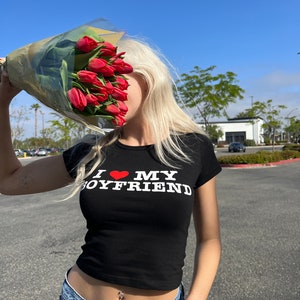 May include: A woman wearing a black crop top with the text "I love my boyfriend" in white letters and a red heart. She is holding a bouquet of red tulips in front of her face.