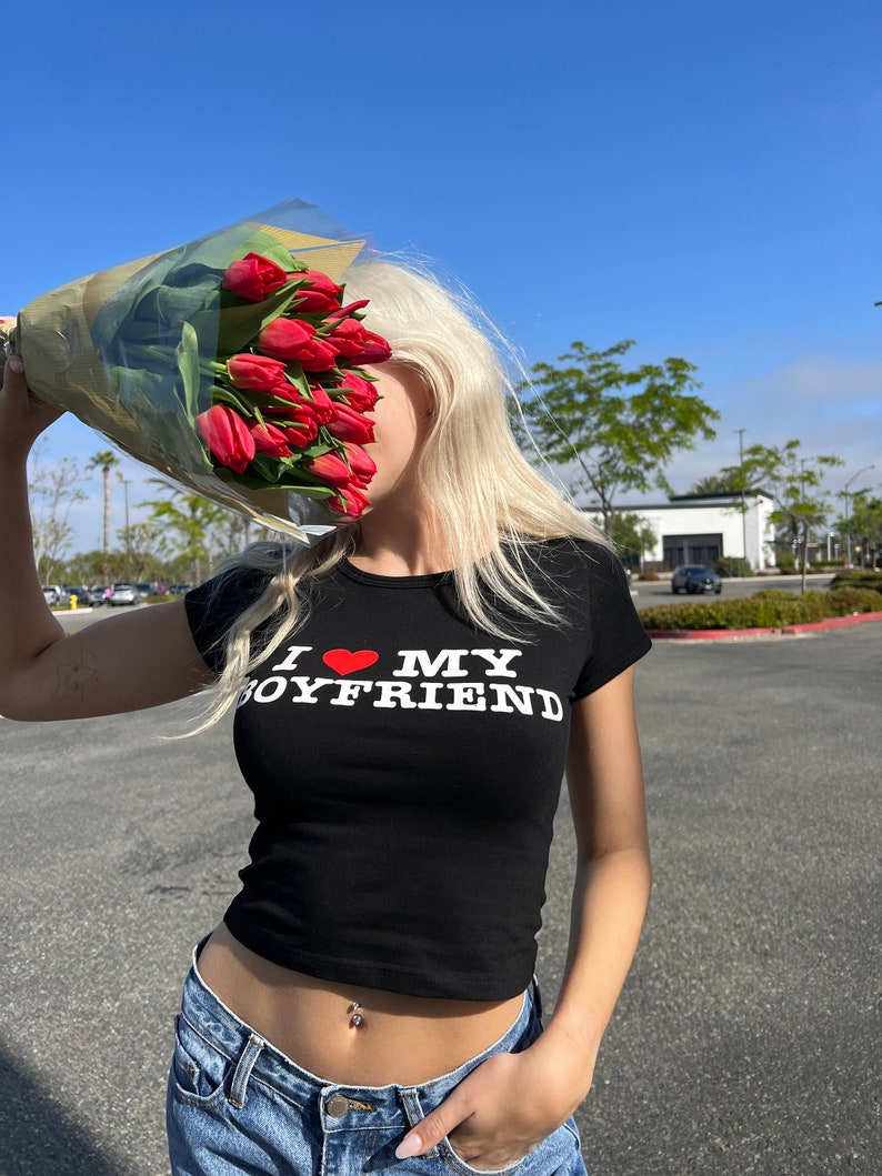 May include: A woman wearing a black crop top with the text "I love my boyfriend" in white letters and a red heart. She is holding a bouquet of red tulips in front of her face.