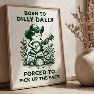 May include: A framed art print featuring a green illustration of a frog playing a banjo while wearing a cowboy hat. The text reads "BORN TO DILLY DALLY" above the frog and "FORCED TO PICK UP THE PACE" below. The print is in a light wood frame.