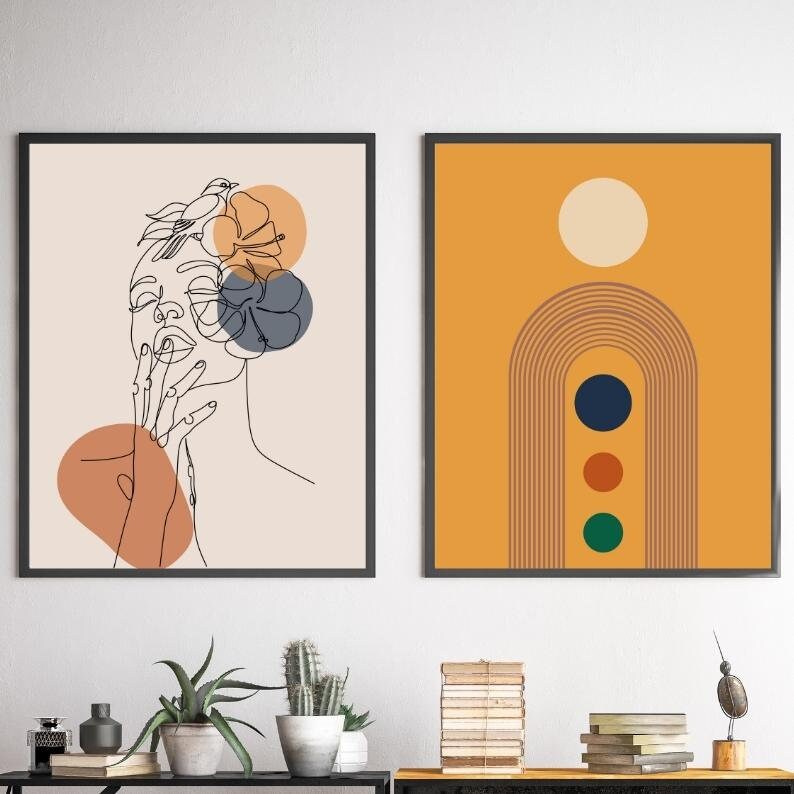 May include: Two framed abstract art prints. The print on the left features a line drawing of a woman's face with a bird and flowers. The print on the right features a rainbow arch with three circles in a yellow background.