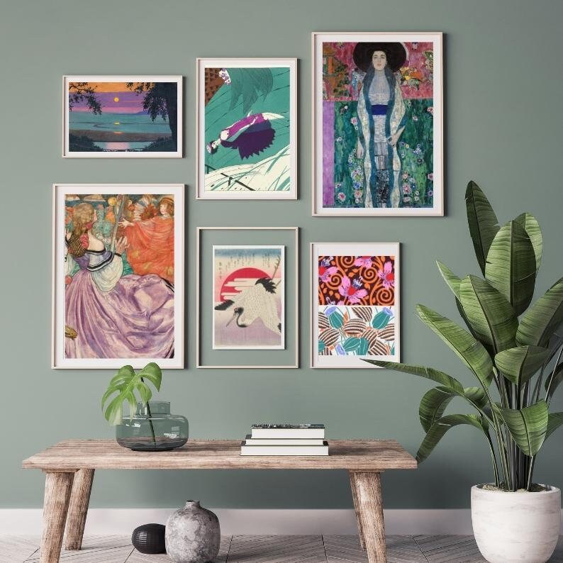 May include: A wall gallery featuring six framed prints. The prints are a mix of abstract and figurative art. The prints are arranged in a grid pattern on a light green wall. A wooden table with a vase of greenery sits in front of the wall. A potted plant with large green leaves is on the right side of the image.