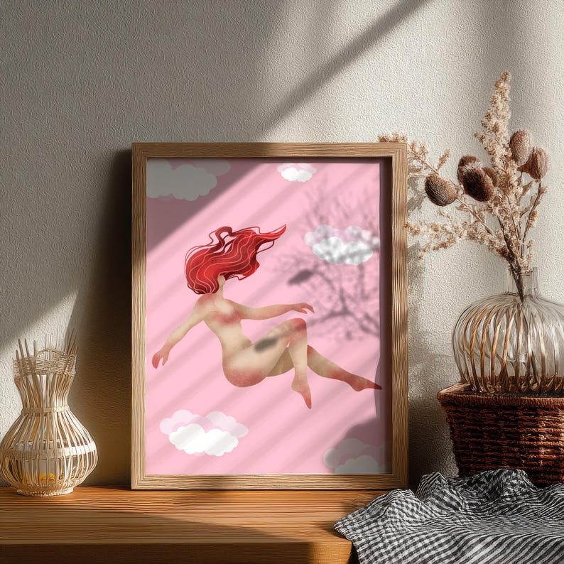 May include: A framed art print featuring a nude figure with flowing red hair floating among white clouds against a pink background. The artwork is in a wooden frame and is displayed on a wooden surface.