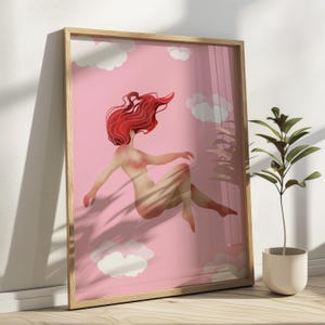 May include: A framed art print featuring a nude figure with flowing red hair floating in a pink sky with white clouds. The artwork is in a wooden frame and leans against a wall. A small potted plant is in the foreground.