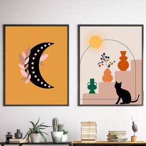 May include: Two framed prints with abstract designs. The first print features a black crescent moon with pink leaves and white polka dots on a yellow background. The second print features a black cat sitting on a pink staircase with a green vase, an orange vase, and a yellow vase with a blue and green plant on a beige background.