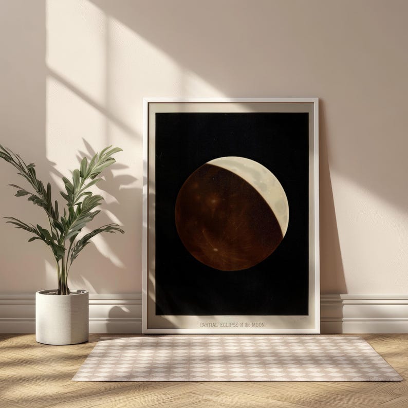 Op de afbeelding: Een ingelijste print van een gedeeltelijke maansverduistering. Het kunstwerk toont een grote, cirkelvormige maan tegen een zwarte achtergrond. De maan is verdeeld, met een heldere, witte halve maan en een donker, roodbruin deel. De tekst "PARTIAL ECLIPSE of the MOON" staat onderaan.